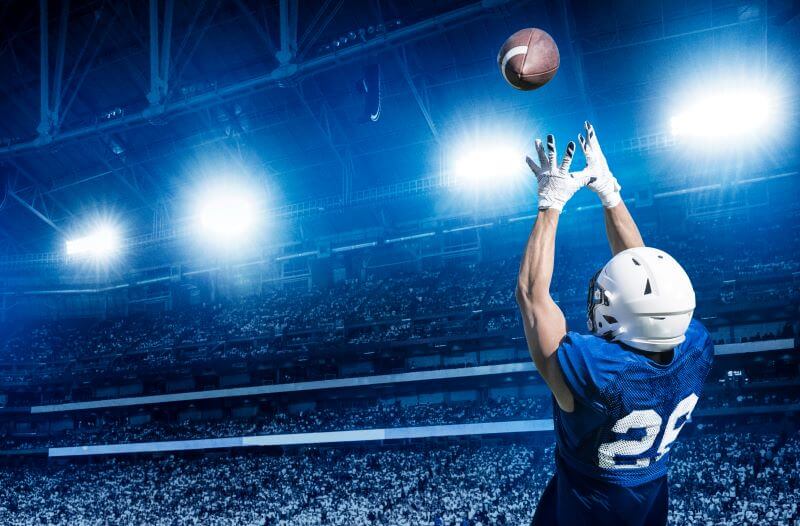 A football player wearing a blue jersey with the number 26 and a white helmet reaching up to catch a football under bright stadium lights. The background shows a large crowd in a packed stadium, emphasizing the intensity and excitement of a game under the night sky.