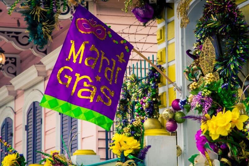 A colorful Mardi Gras flag hanging outside a decorated building, featuring purple, green, and gold colors with the text 'Mardi Gras' and a mask graphic. The building is adorned with festive decorations such as garlands, ribbons, and flowers in vibrant colors, capturing the lively and celebratory spirit of the Mardi Gras festival.