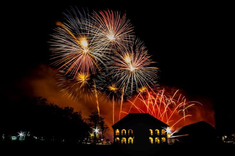 Bright and colorful fireworks exploding in the night sky above a large building with illuminated windows. The scene is set against a dark background, with trees and additional buildings partially visible, creating a festive and celebratory atmosphere.