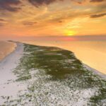 A stunning aerial view of a narrow stretch of sandy beach extending into the distance with a calm ocean on both sides. The sunset casts a warm golden glow across the sky, with hues of orange, pink, and purple blending together. The shoreline curves gently along the water, and sparse greenery dots the sand. The peaceful scene captures the beauty of a coastal landscape during a serene sunset.