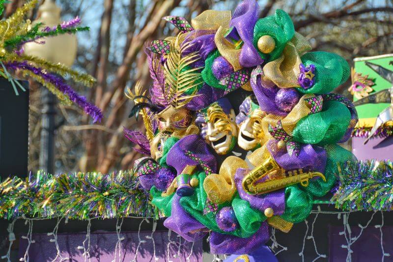 A vibrant Mardi Gras wreath decorated with purple, green, and gold mesh ribbons, beads, and festive masks with feathers and glitter. The wreath includes decorative elements like a small saxophone, showcasing a lively and colorful theme. It is displayed on a float or a structure adorned with garlands and other Mardi Gras decorations, capturing the festive spirit of the celebration.